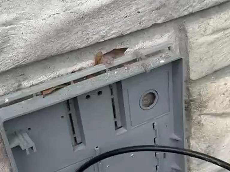 An exterior dryer vent hood with its cover off on a stone wall, a cable entering from the right