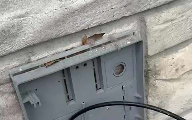 An exterior dryer vent hood with its cover off on a stone wall, a cable entering from the right